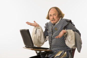 William Shakespeare in period clothing sitting in school desk with laptop computer.